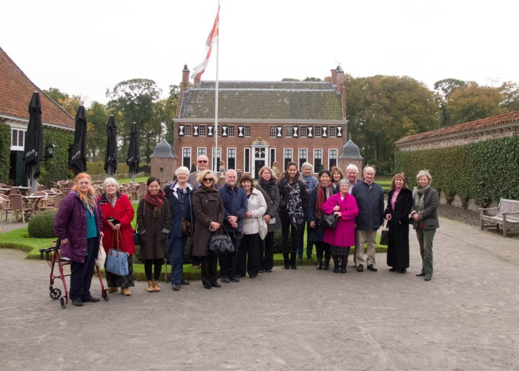 18 attendees of the OCS Visit to Dutch Collections standing by a flagpole outside a symmetrical house with building to each side, all smiling at the camera